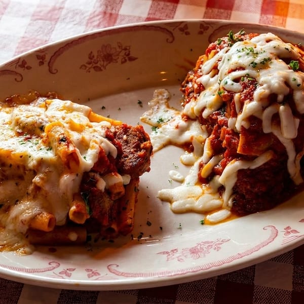 Baked pasta and a large meatball covered in red sauce and melted cheese on a checkered tablecloth.