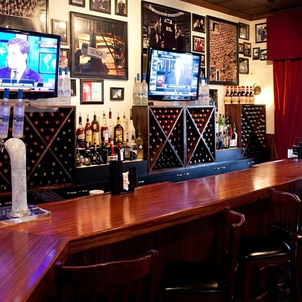 Restaurant bar with dark wood counter, liquor bottles, wine racks, two TVs, and red curtains.