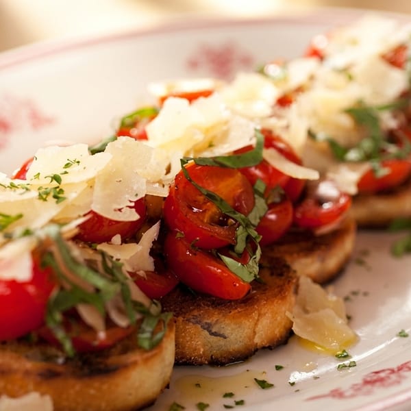 Toasted bruschetta topped with fresh cherry tomatoes, basil, and shaved Parmesan cheese on a white plate.