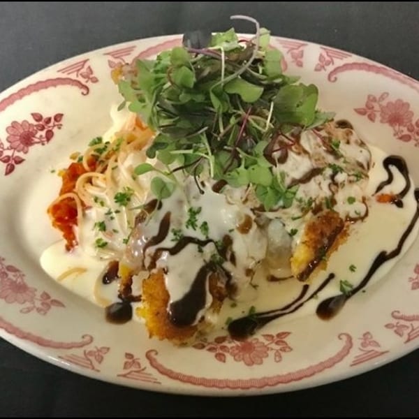 Crispy fried chicken cutlet topped with creamy sauce, balsamic glaze, pasta, and microgreens on a decorative plate.