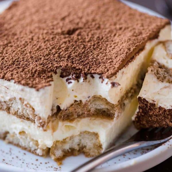 A large slice of tiramisu with a fork taking a piece, dusted with cocoa powder.