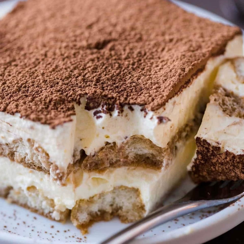 A large slice of tiramisu with a fork taking a piece, dusted with cocoa powder.